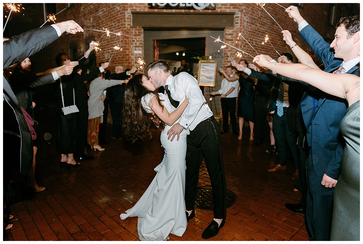 groom dips bride and kisses her as guests hold sparklers by The Hazel Club