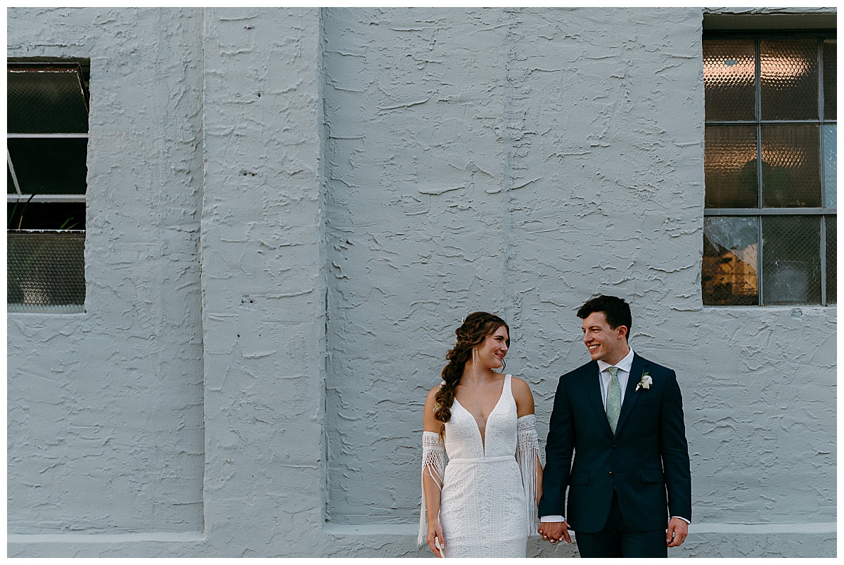 bride and groom hold hands and look at each other in front of building by The Hazel Club